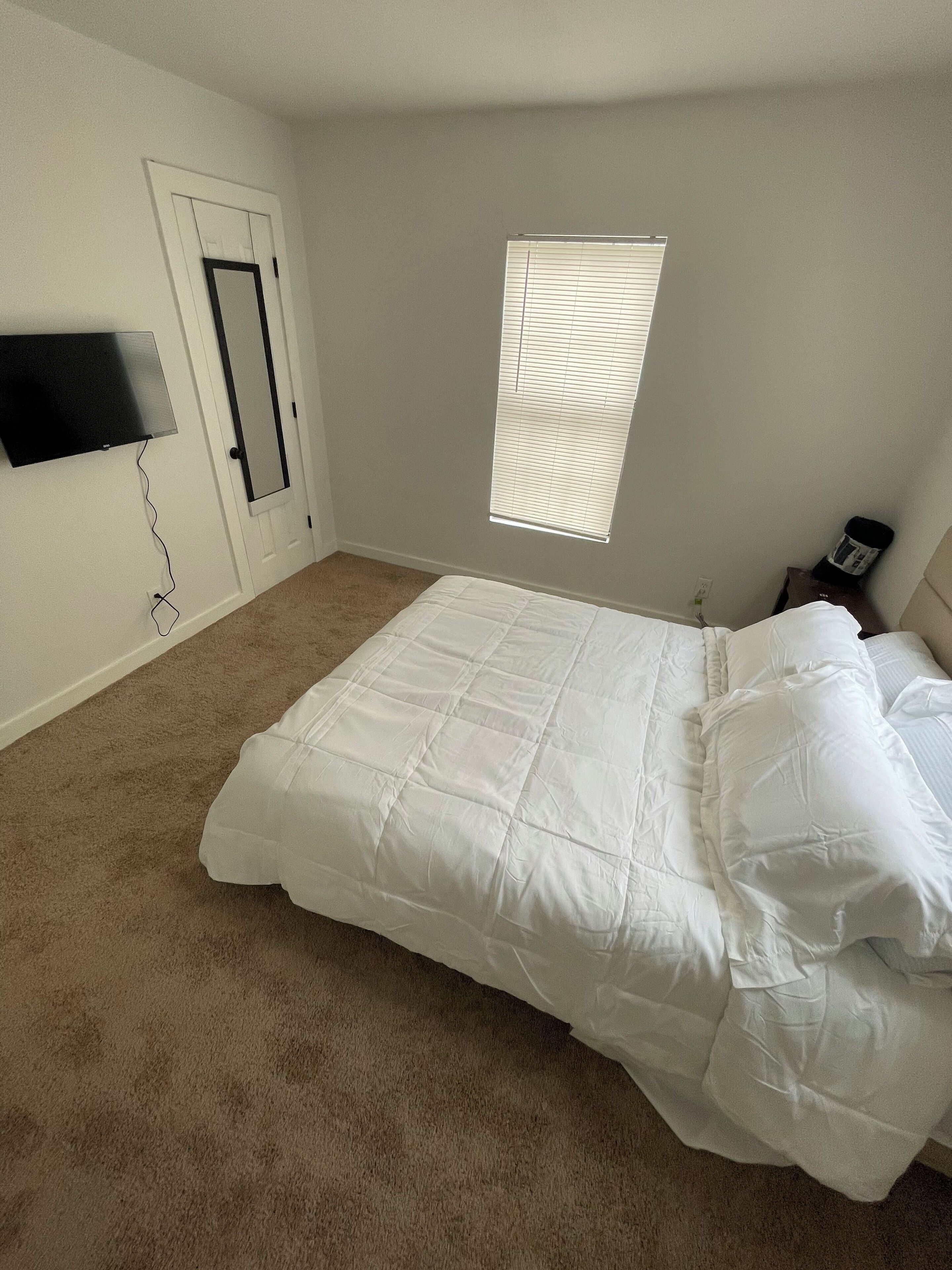 Ein Schlafzimmer mit einem Bett, einer Tür, einem an der Wand montierten Fernseher und einem Fenster mit Jalousien.