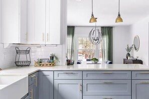 A modern kitchen with white cabinets, a grey countertop, and gold handles. There are two pendant lights, a window with green curtains, and a small plant on the counter. - Sleeps 12, Firepit, near Downtown/SU (Syracuse)