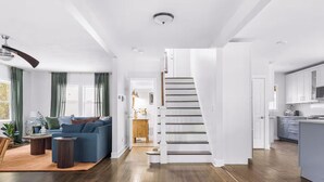 A modern interior with a staircase leading to a second floor, a living area with a blue sofa, wooden flooring, and a kitchen with white cabinets. - Sleeps 12, Firepit, near Downtown/SU (Syracuse)