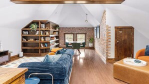 A loft-style room with a high ceiling, exposed wooden beams, a brick wall, a wooden bookshelf, a blue sofa, a dining table with chairs, and a wooden door. - Sleeps 12, Firepit, near Downtown/SU (Syracuse)