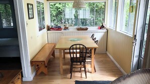 Een gezellige keuken met een eettafel, stoelen en een bankje. Er staan manden op een plank, een hanglamp en een raam met uitzicht op bomen.