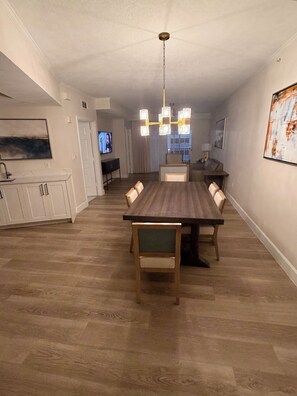 Un espace repas avec une longue table en bois, des chaises et un lustre. On y trouve également un coin cuisine avec des placards et un téléviseur en arrière-plan.