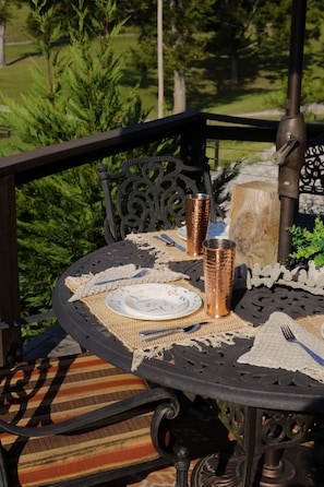 A round outdoor table with a metal frame, surrounded by a woven placemat, a plate, a fork, a knife, and two copper-colored cups.