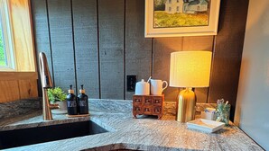 A kitchen with a granite countertop, wooden cabinets, a sink, a faucet, a small cabinet with kitchen items, a towel hanging on the cabinet door, and a painting on the wall. - Working Horse Ranch Retreat – 2 Bedrooms, Fire Pit, 20 Min to Downtown Nashville (Goodlettsville)