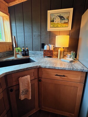 Une cuisine avec un comptoir en granit, des armoires en bois, un évier, un robinet, un petit meuble contenant des ustensiles de cuisine, une serviette accrochée à la porte de l’armoire et un tableau au mur.