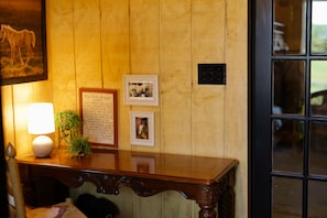 A wooden wall with framed pictures and a plaque, a table with a lamp and a plant, and a door with a window reflection.