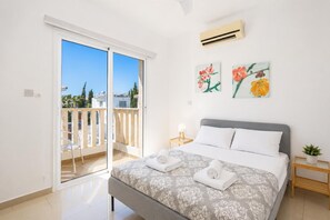 A bedroom with a bed, a nightstand, a potted plant, and a balcony with a railing. - Villa Chromata - Three Bedroom Villa, Sleeps 7 (Pernera)