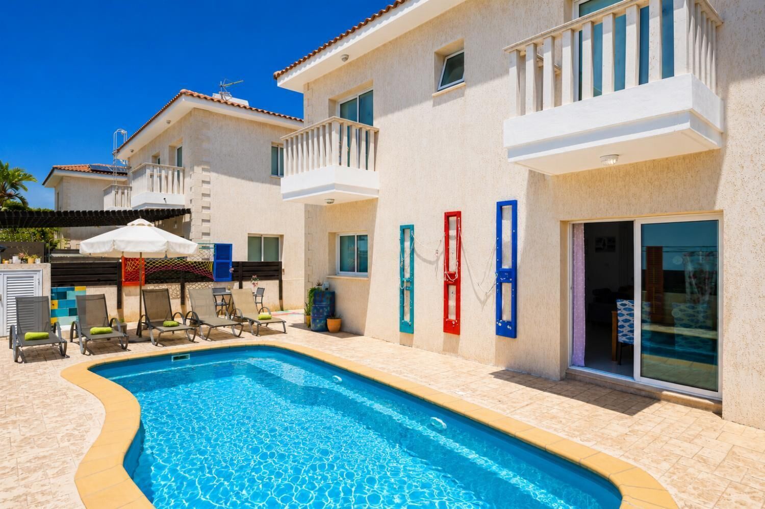 A swimming pool with lounge chairs, a covered seating area, and a building with balconies and sliding doors.