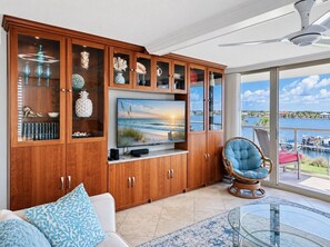 Ein Wohnzimmer mit großem Fernseher, Holzkredenz, Korbsessel und Blick auf das Wasser.