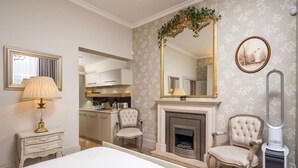 Une chambre à coucher avec un grand lit, une cheminée, deux fauteuils, un miroir et une table de chevet avec une lampe.