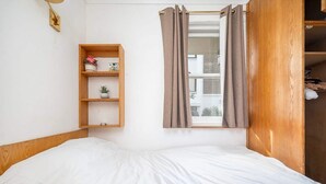 Un lit simple avec une tête de lit en bois, une petite étagère en bois ornée d’objets décoratifs et une fenêtre avec des rideaux.