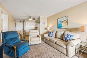 A living room with a blue armchair, beige sofa, and a painting of a beach scene. - Ocean & Inlet Views | Cherry Grove Condo (North Myrtle Beach)