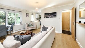 Een moderne woonkamer met een bank, salontafel en een eethoek met stoelen. Achterin is een keuken, een houten deur en een groot raam met uitzicht op groen.