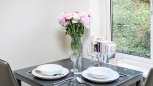 Een eettafel met borden, bestek, een vaas met bloemen en boeken bij het raam.