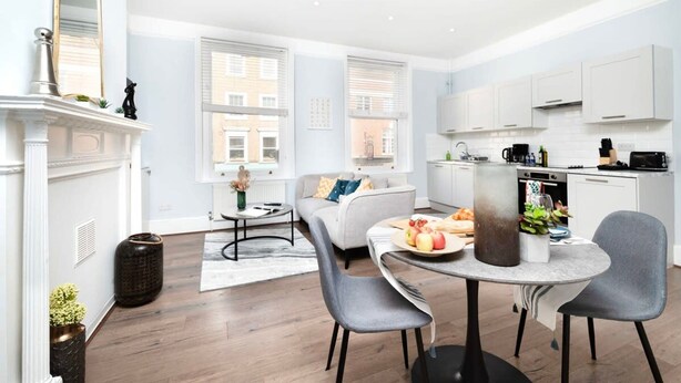 A modern living room with a fireplace, a dining table with chairs, a sofa, and a kitchen area with cabinets and appliances. - Serene One-Bedroom Retreat in Fitzrovia (London)