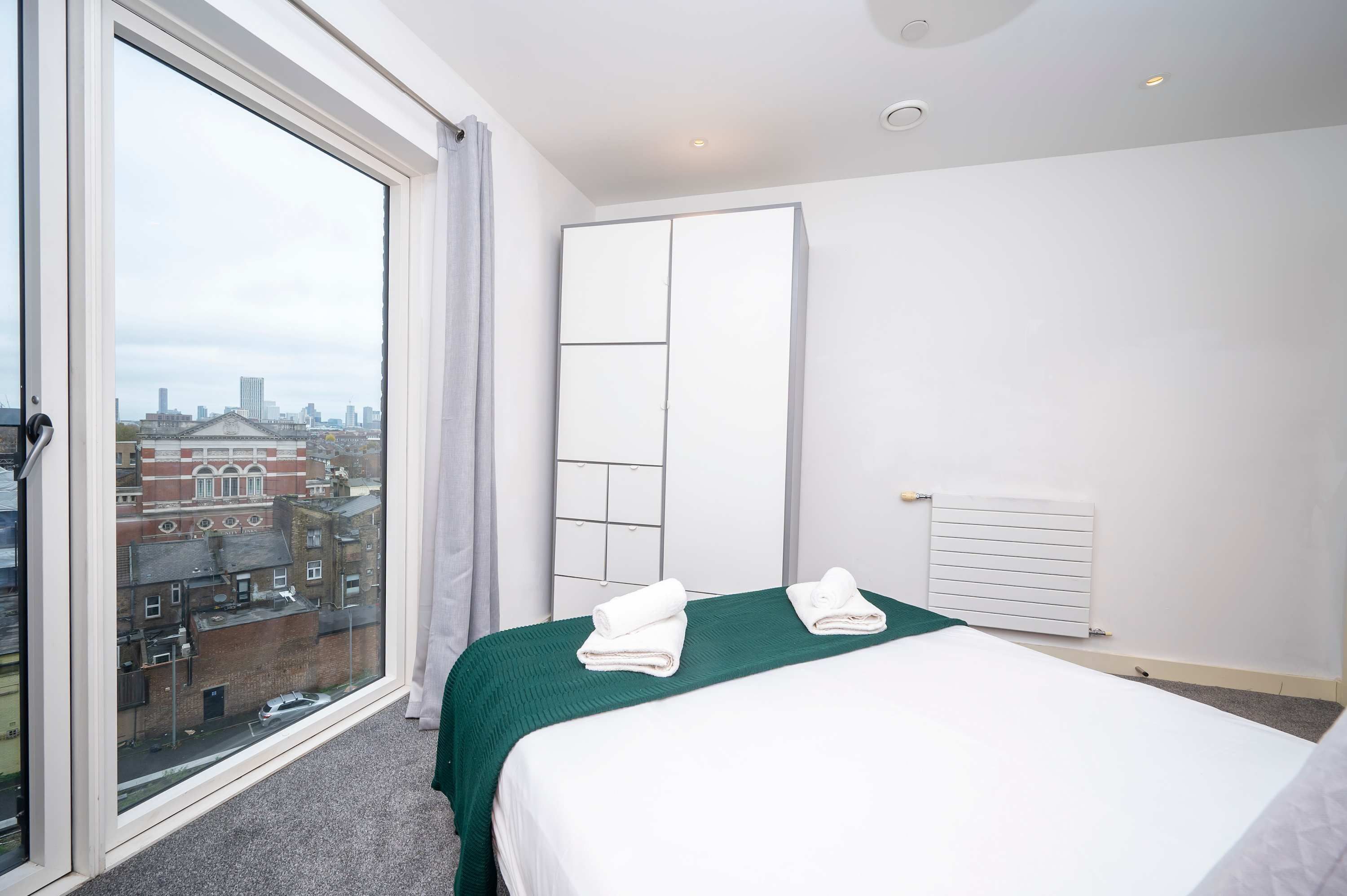 Une chambre d’hôtel avec une grande fenêtre, un lit avec une literie verte, un radiateur et une armoire.