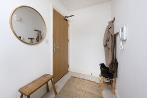 Um corredor com um banco de madeira, um espelho redondo, uma porta, um cabideiro com casacos e um sapateiro.