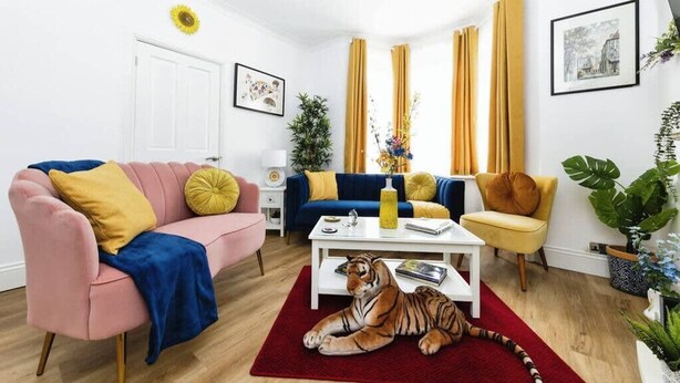 En stue med en lyserød sofa, en blå sofa, en gul lænestol og en tigerfigur på et rødt tæppe.