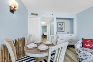A dining area with a round table set for four, white chairs, and a wall-mounted light fixture. - Compass Cove 268: Oceanfront (Myrtle Beach)