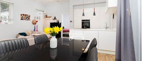 Una sala moderna con una mesa de comedor negra, un sofá y una mesa de centro con flores amarillas. Se aprecia una zona de cocina con estufa y lavaplatos.