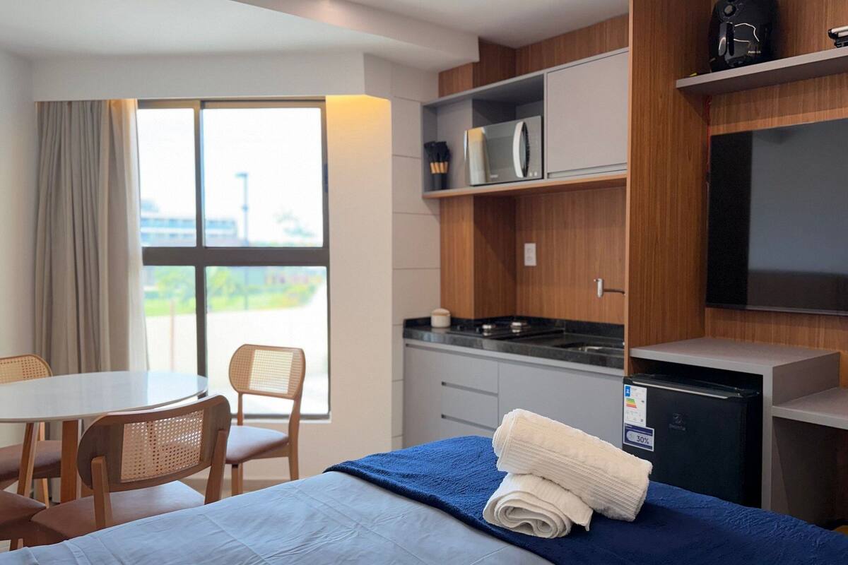 A hotel room with a bed, a desk, a chair, a small kitchenette, and a TV.
