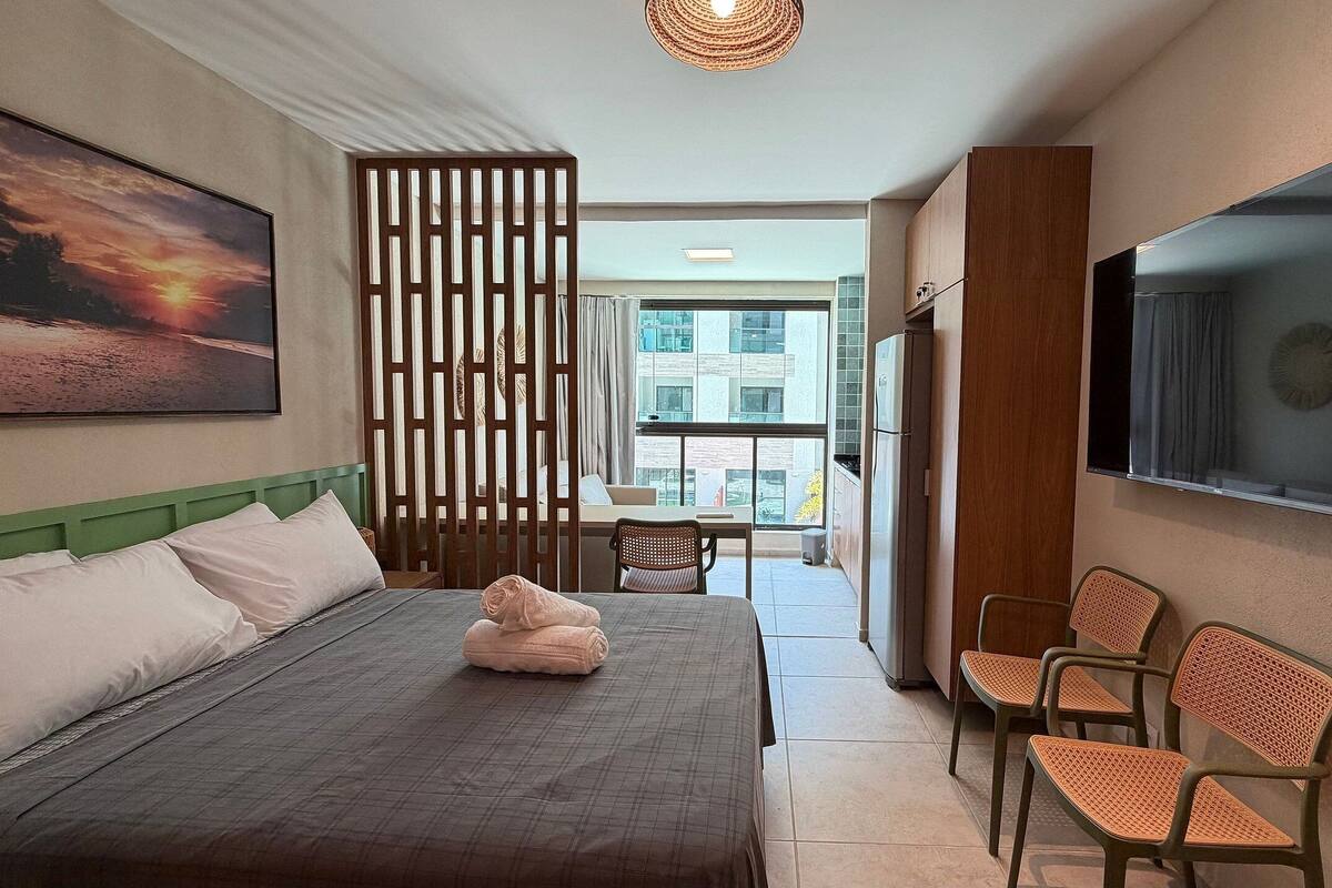 A hotel room with a bed, a desk, chairs, a TV, and a view of the city.
