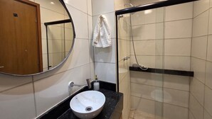 A bathroom with a round mirror, a sink, a towel rack, and a glass-enclosed shower. - Max Carneiros (Tamandaré)
