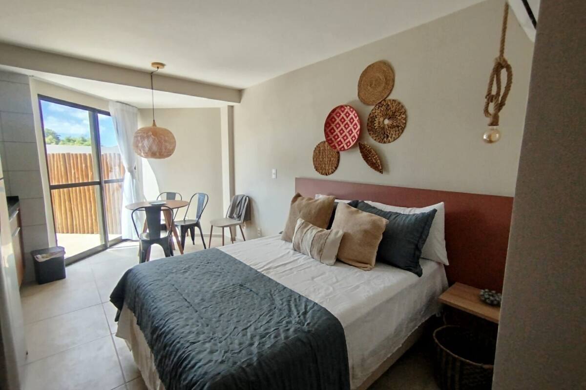 A bedroom with a bed, pillows, a headboard, a wall with decorative items, a dining table with chairs, and a view through a sliding door.