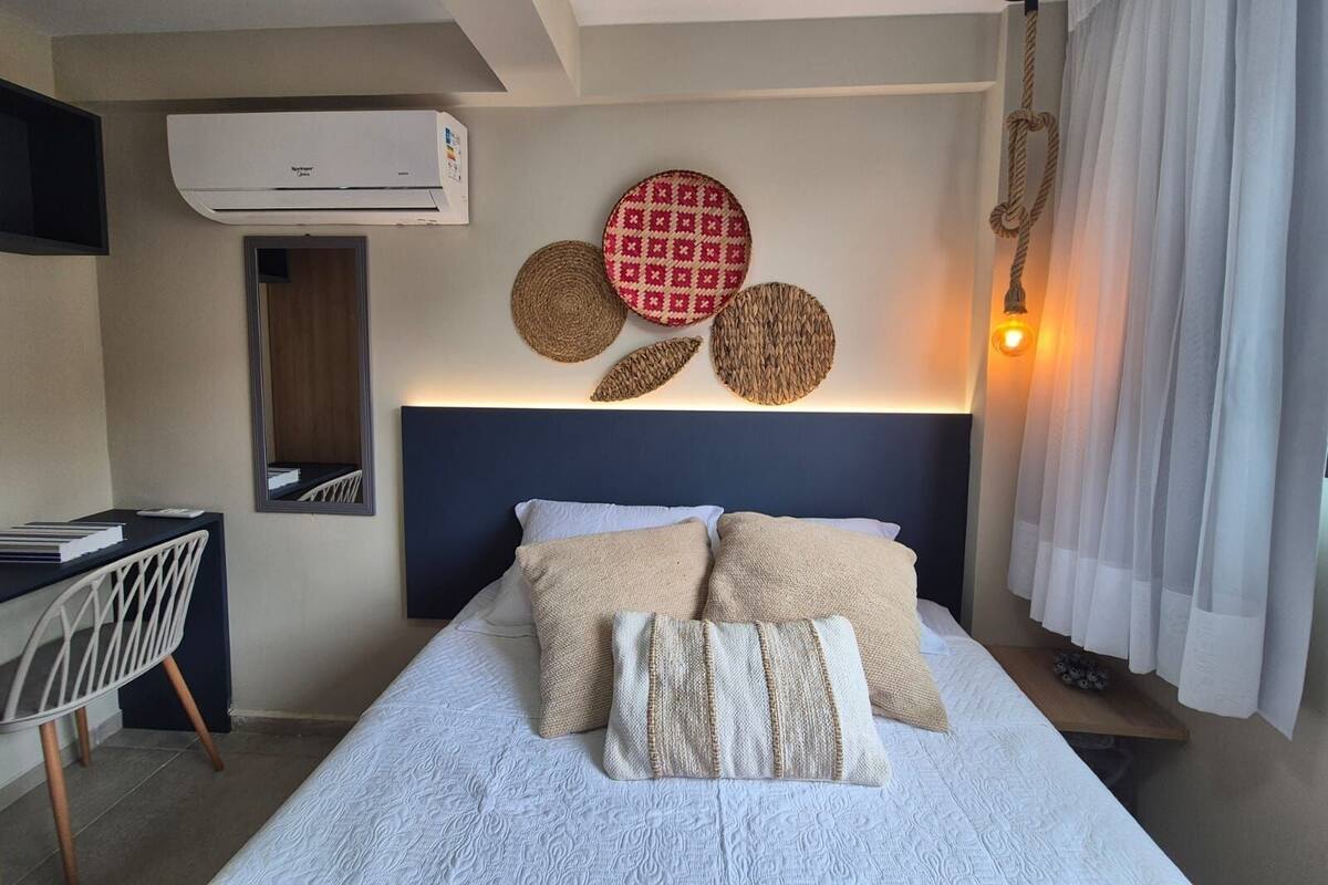 A hotel room with a bed, a desk, a mirror, and a wall-mounted air conditioner.