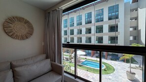 A modern living room with a large window, a circular wall decoration, and a view of a building with multiple windows and a courtyard with a pool. - Max Carneiros (Tamandaré)