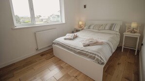 A bedroom with a large window, a bed with white bedding, two bedside tables with lamps, and a radiator. - Spacious Home Near Porthtowan Beach (Porthtowan)