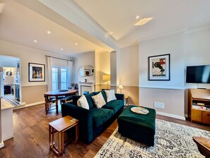 A modern living room with a green sofa, a coffee table, and a TV. There is a dining area with a table and chairs, and a kitchen visible in the background. - Charming Two-Bedroom Home in London (Greater London)