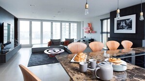 A modern living room with a large window, a sofa, a coffee table, and a dining area with a marble table and chairs. - Stylish Penthouse with London Views (London)