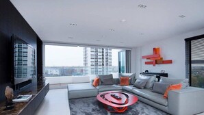 A modern living room with a grey sofa, a red coffee table, and a large window offering a city view. - Stylish Penthouse with London Views (London)