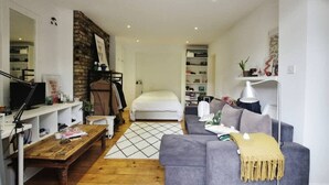 A modern living room with a sofa, coffee table, and a TV. There is a bed in the background and a brick wall. - Cozy Studio with Private Garden in London (London)
