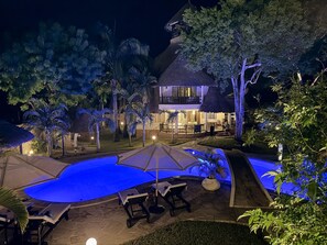 Vista nocturna de un resort con piscina, sillas de descanso y un edificio de techo de paja.