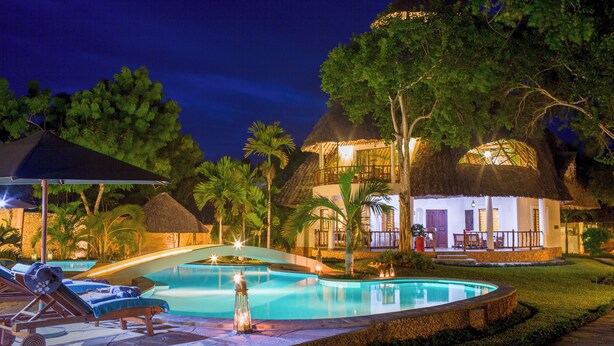 Un espace piscine d’un complexe hôtelier, aménagé pour la nuit, avec des chaises longues, une piscine et des structures à toits de chaume.