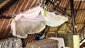 Une pièce avec un toit de chaume, un lit recouvert d’une courtepointe à motifs léopard, une table avec une nappe à rayures zébrées et une moustiquaire suspendue.