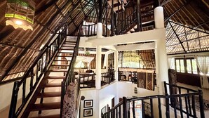 Un escalier en bois à plusieurs niveaux, surmonté d’un toit de chaume, avec une lanterne suspendue, et donnant sur une pièce dont le sol est décoré de motifs.