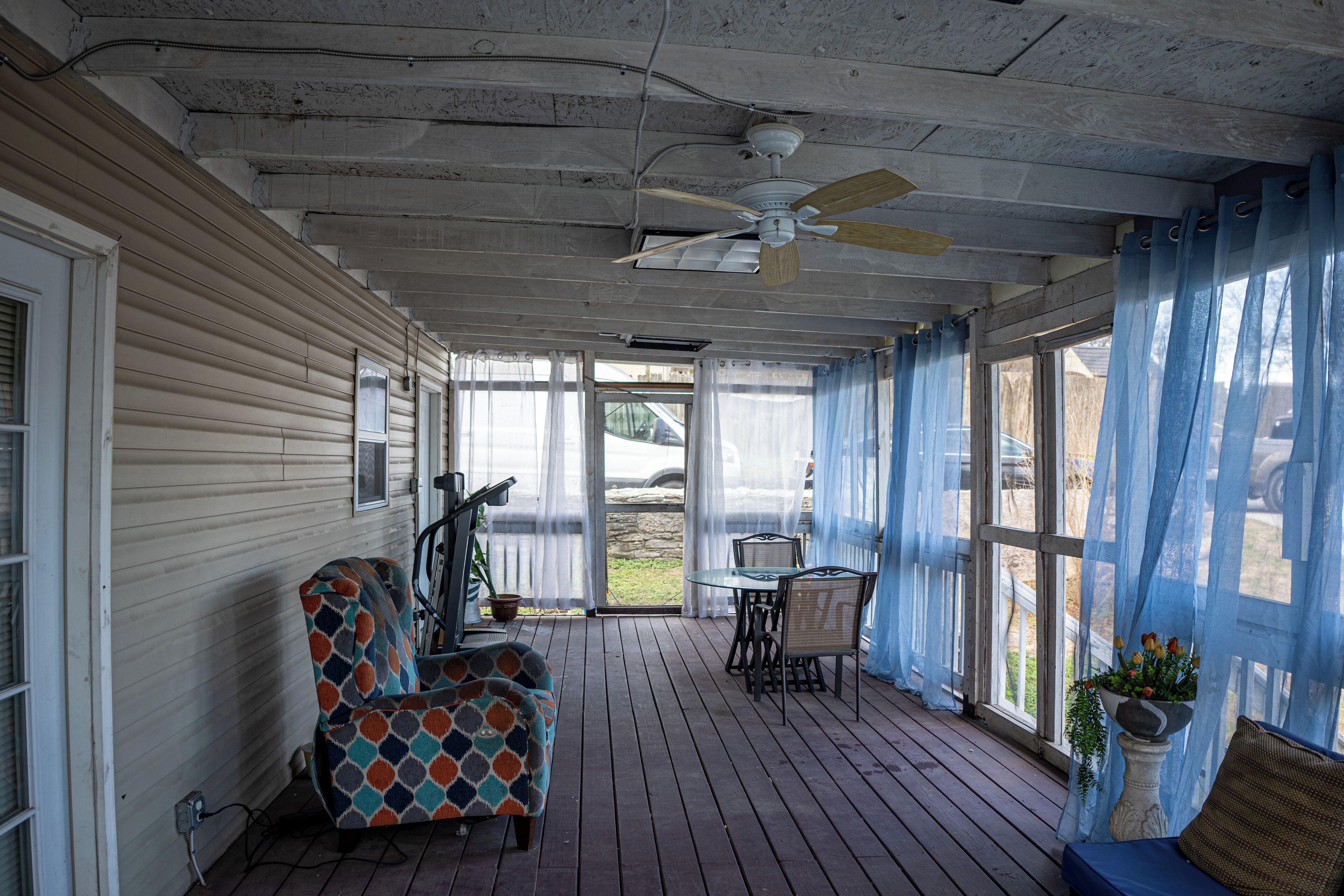 A covered porch with a ceiling fan, a dining table with chairs, a patterned armchair, and a potted plant.