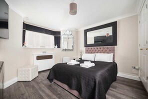 Ein modernes Schlafzimmer mit einem großen Bett, einem Spiegel, einer weißen Kommode und einem kleinen Hocker.
