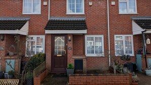 A two-story brick house with a front door, two windows on the ground floor, and a small garden area with potted plants and a wooden fence. - Comfortable 2-Bedroom house with free parking (Greater London)
