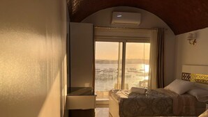 A hotel room with two beds, a large window with a view of a body of water, an air conditioning unit, and a small table with a lamp. - Wanas kato  (Aswan)