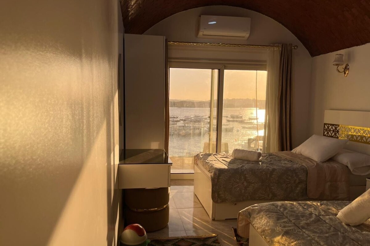 Quarto de hotel com duas camas, uma janela grande com vista para um corpo de água, um ar condicionado e uma pequena mesa com uma luminária.