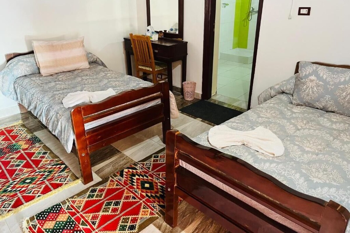 Quarto com duas camas de madeira, uma escrivaninha, uma cadeira e um espaço com chuveiro.