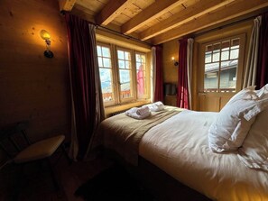 Ein Zimmer mit einem Bett, zwei mit Vorhängen versehenen Fenstern, einem Sessel und Wandleuchten.