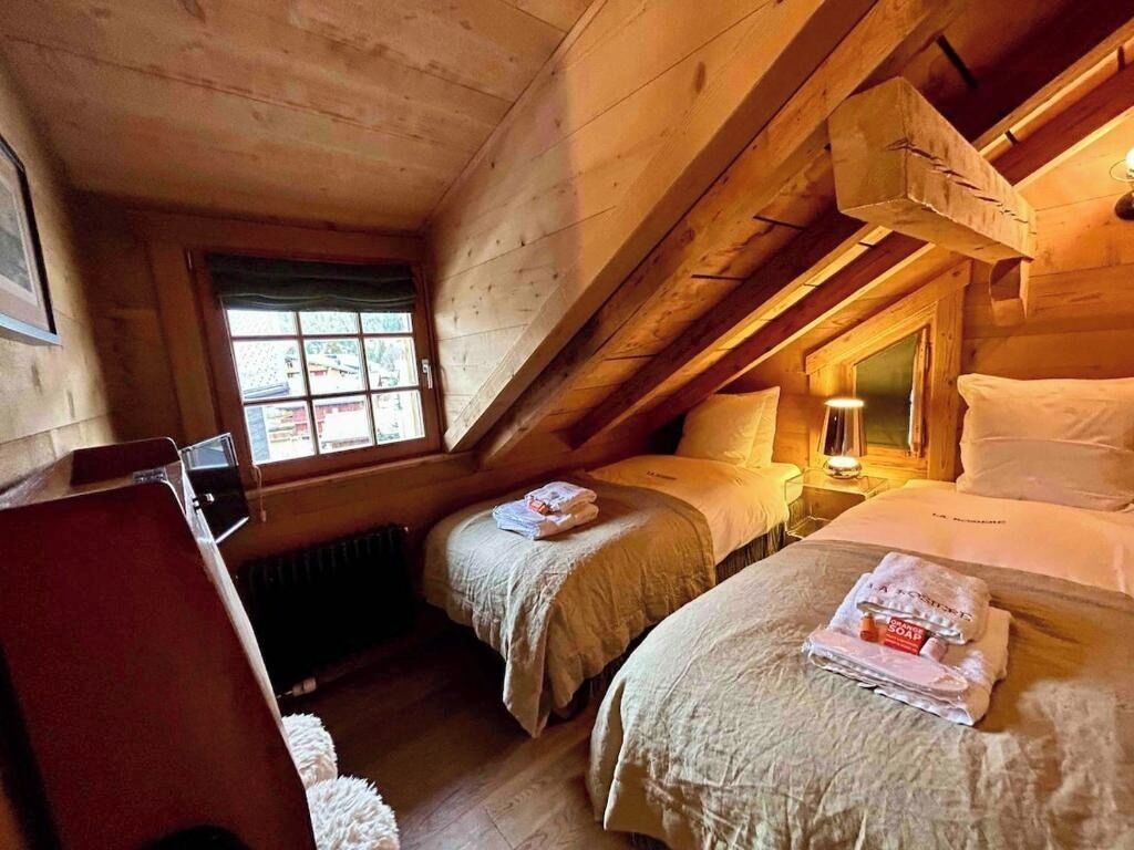 A cozy attic bedroom with two beds, a window, and a wooden ceiling.