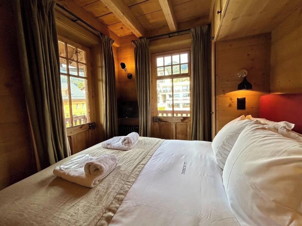 A hotel room with a bed, two windows, a lamp, and a view of the outside.