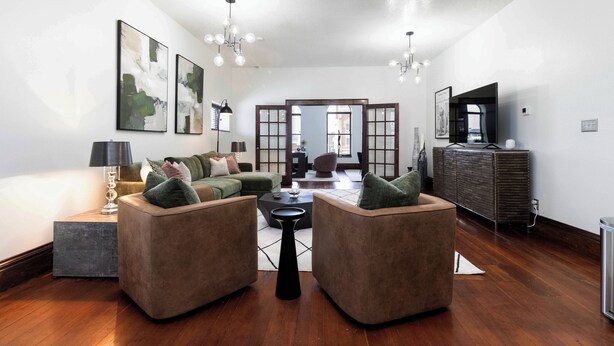 Een moderne woonkamer met een hoekbank, een salontafel en een televisie op een meubel. Op de muur hangen abstracte schilderijen en van het plafond hangt een kroonluchter.