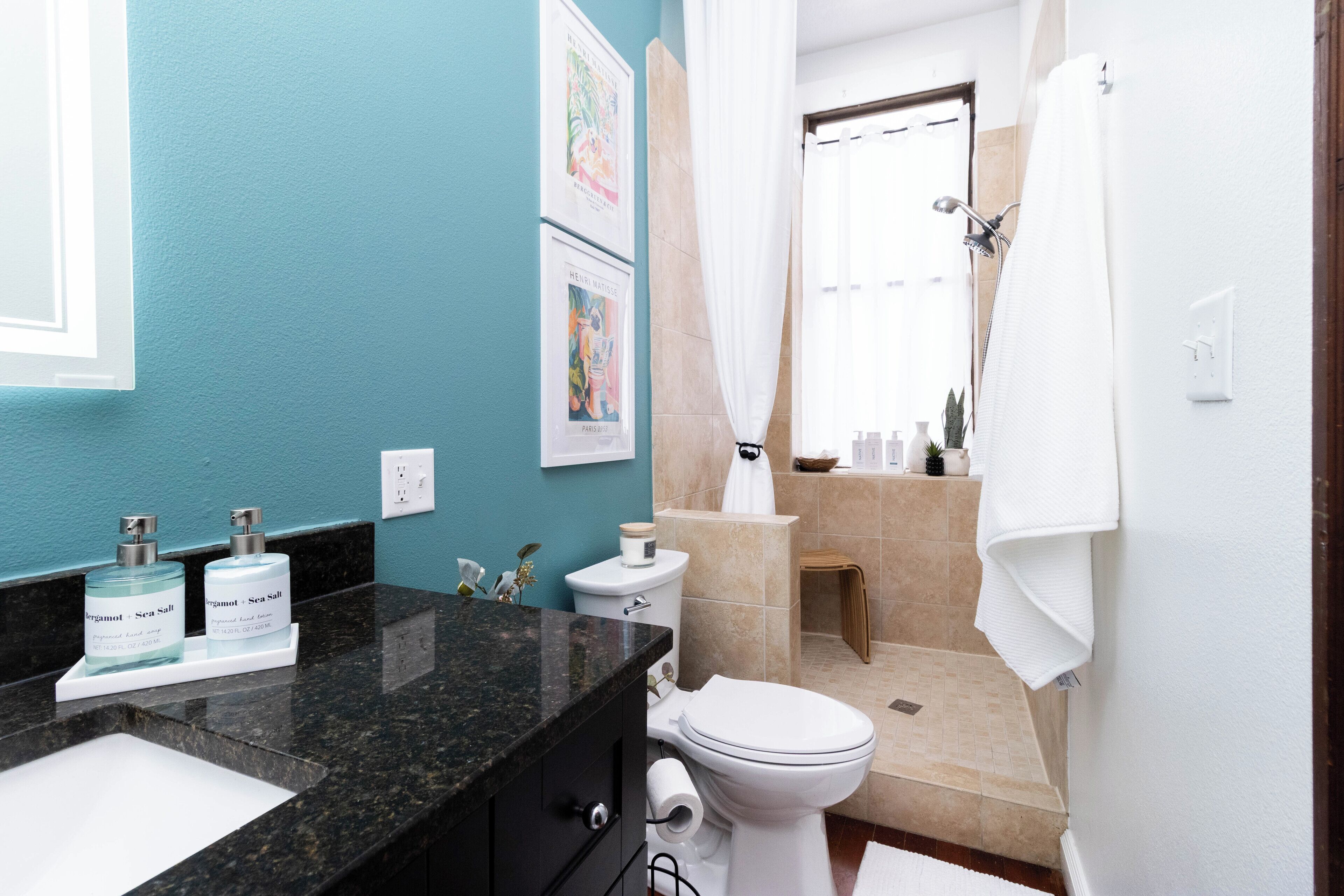 Une salle de bain avec un plan de toilette en granit noir, un lavabo blanc et des toilettes. On peut y voir deux bouteilles de sel marin posées sur le plan de toilette.
